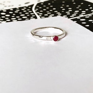 May include: A silver ring with a hammered finish and a small, round, red gemstone. The ring is set against a white background with a black and white pattern in the background.