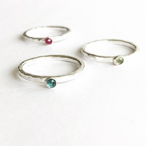 May include: Three silver rings with hammered bands, each featuring a different coloured gemstone: red, green, and teal. The rings are set against a white background, showcasing their simple elegance and delicate design.