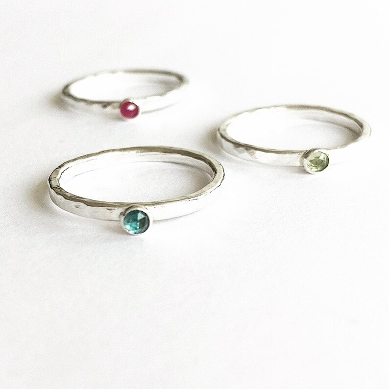 May include: Three silver rings with hammered bands, each featuring a different coloured gemstone: red, green, and teal. The rings are set against a white background, showcasing their simple elegance and delicate design.