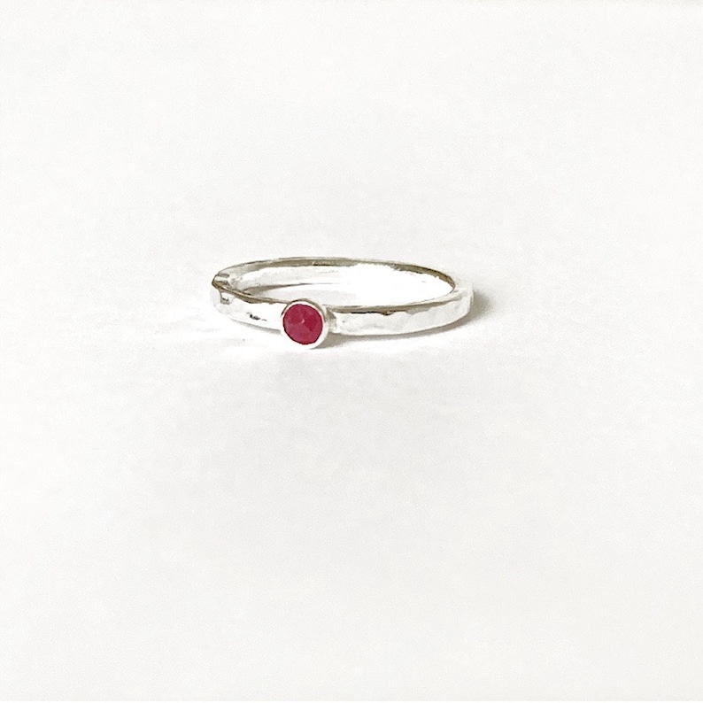 May include: A silver ring with a hammered finish, featuring a small, round, red gemstone. The ring's design is simple, set against a white backdrop, emphasizing the gemstone's colour and the ring's texture.