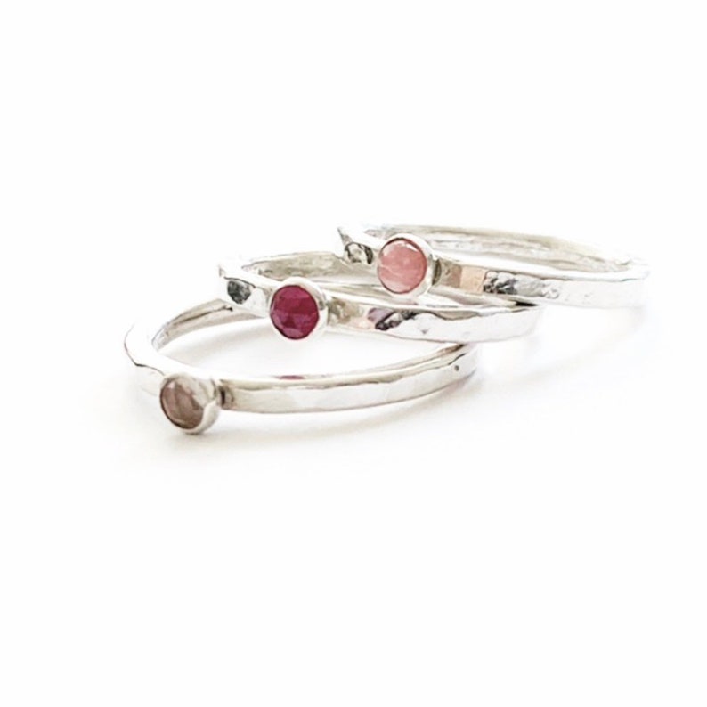 May include: Three silver rings with textured bands, each set with a small, round gemstone. The gemstones are in shades of pink and red. The rings are stacked on a white surface.