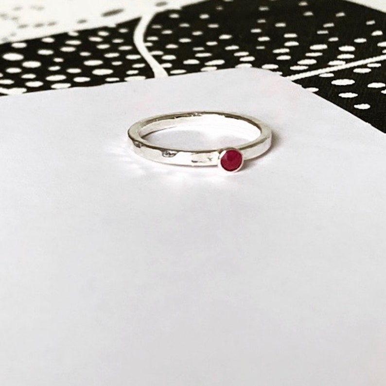 May include: A silver ring with a hammered finish and a small, round, red gemstone. The ring is set against a white background with a black and white pattern in the background.