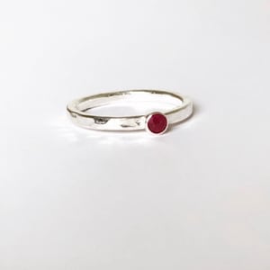May include: A silver ring with a hammered finish, featuring a small, round, red gemstone. The ring is displayed on a white backdrop, accentuating the contrast between the metal and the vibrant stone. A simple and elegant piece of jewellery.