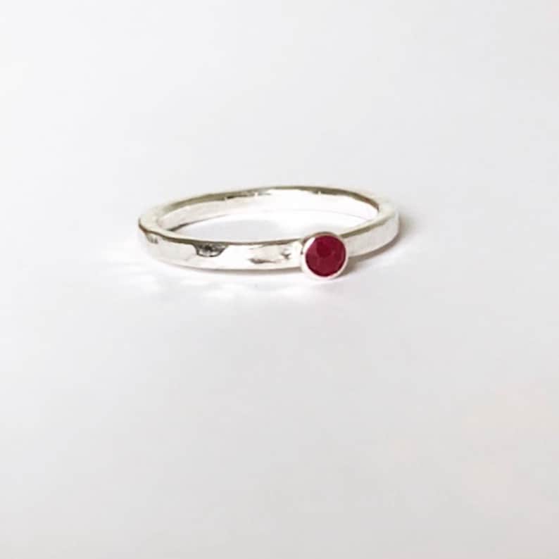 May include: A silver ring with a hammered finish, featuring a small, round, red gemstone. The ring is displayed on a white backdrop, accentuating the contrast between the metal and the vibrant stone. A simple and elegant piece of jewellery.