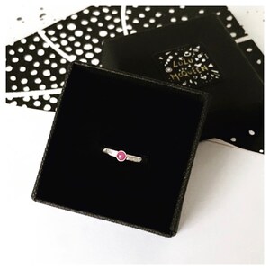 May include: A silver ring with a small, round, red gemstone presented in a black jewellery box. The ring is set against a black and white patterned card and a black box with the text "Lulu McQueen".