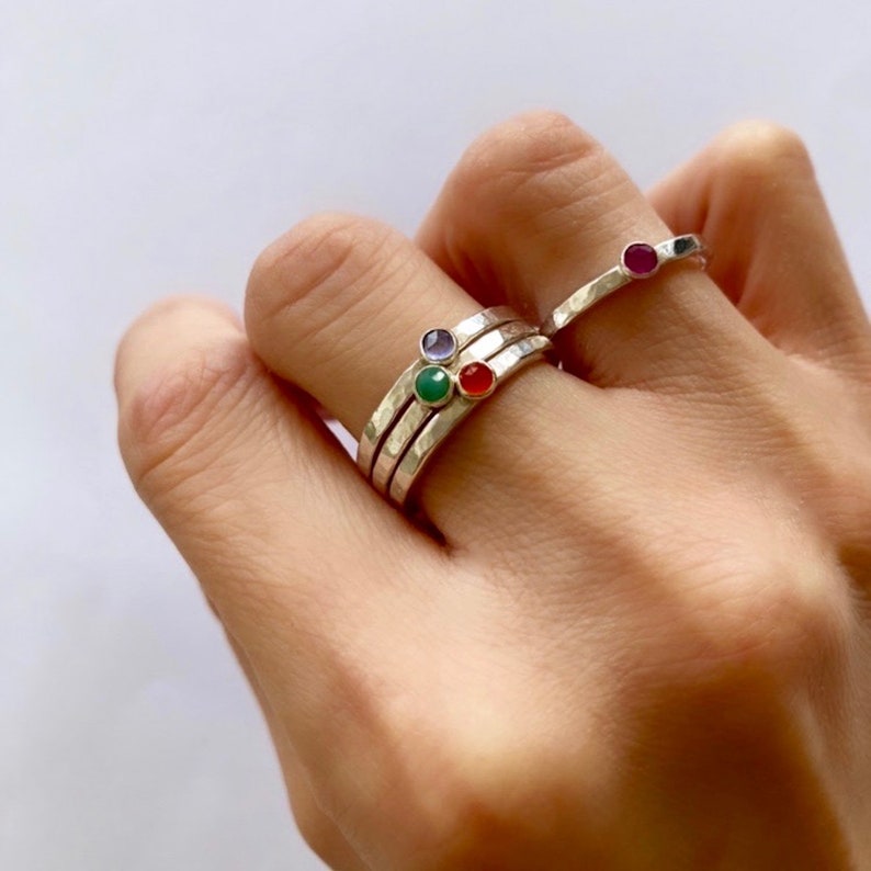 May include: A hand displays a collection of silver rings, each with a hammered texture and a small, colourful gemstone. The gemstones are purple, green, red, and ruby. The rings are stacked on the fingers.