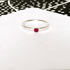 May include: A silver ring with a hammered finish, featuring a small, round, red gemstone. The ring is set against a white backdrop, with a black and white patterned fabric in the upper left corner.