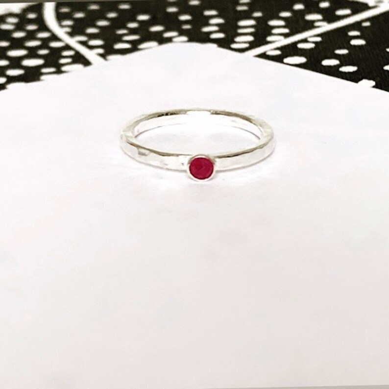 May include: A silver ring with a hammered finish, featuring a small, round, red gemstone. The ring is set against a white backdrop, with a black and white patterned fabric in the upper left corner.