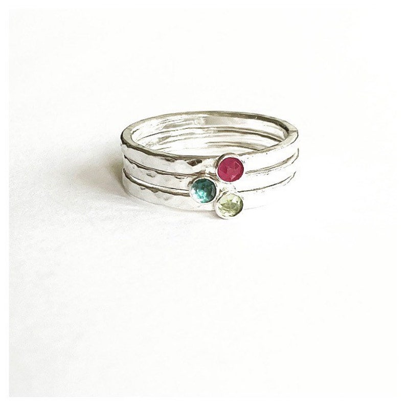 May include: Four silver rings with a hammered finish, stacked together. Each ring features a small, round gemstone in red, teal, and light green. The rings are presented on a white backdrop.