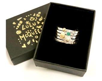 May include: A silver ring with multiple bands, featuring a green gemstone and gold accents. The ring is in a black box with the text "Lulu McQueen" on the lid.
