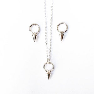 May include: A silver jewelry set featuring a necklace and matching earrings. The necklace has a delicate chain with a ring pendant and a cone-shaped charm. The earrings also have a ring and cone design. The jewelry is set against a white background.