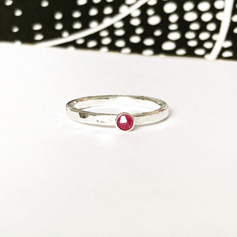 May include: A silver ring with a hammered finish, featuring a small, round, red gemstone. The ring is set against a white backdrop, with a black and white patterned design visible in the background.