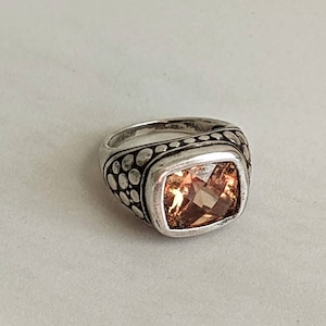 May include: A silver ring with a square, peach-colored gemstone. The band features a dotted design along the top and sides. The gemstone is faceted, catching the light and adding sparkle to the jewelry.