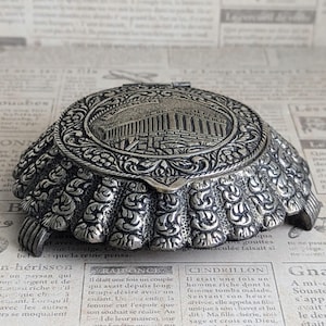 May include: Ornate silver-toned trinket box with a detailed relief of a classical building on the lid. The box has a scalloped, ruffled edge and intricate floral patterns. The box is likely made of metal and is displayed on a vintage newspaper background.