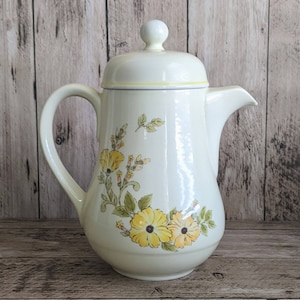 May include: A white ceramic coffee pot with a floral design featuring yellow and green flowers. The pot has a lid with a knob on top.