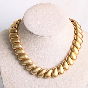 May include: A gold-toned necklace featuring a series of interconnected, smooth, curved, teardrop-shaped metal links. The links are arranged in a continuous, repeating pattern to form a thick, substantial collar-style chain with a matte finish.
