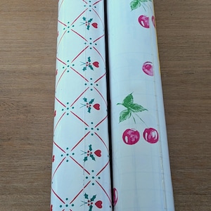 May include: Two rolls of decorative paper. One roll features a white background with a red and green holly and heart pattern. The other roll has a white background with a cherry design. The paper is rolled up.