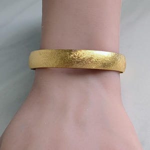 May include: A gold-colored bangle bracelet with a textured surface. The bracelet is wide and has a simple, elegant design. The gold color is bright and shiny, reflecting light.