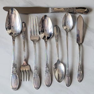 Puede incluir: Un juego de ocho piezas de cubiertos plateados, que incluye un cuchillo para mantequilla, un tenedor y seis cucharas. Los cubiertos presentan un diseño decorativo de vid.