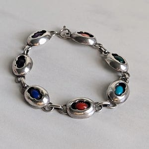 May include: A silver bracelet with a chain link design and six oval-shaped charms. Each charm features a different colored gemstone: purple, pink, green, blue, turquoise, and red.