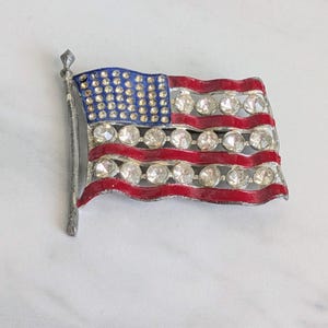May include: A vintage brooch in the shape of the American flag. The flag features a blue field with clear rhinestones, red stripes, and clear rhinestone accents. The flag is mounted on a silver-tone metal pole.