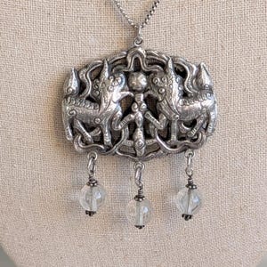 May include: Silver-toned pendant necklace featuring an ornate, rectangular centerpiece with detailed relief carvings of mythical creatures. Three clear, spherical beads dangle below. The necklace hangs on a delicate chain, creating a vintage aesthetic.
