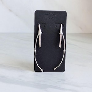 May include: A pair of silver-colored earrings in a unique, branch-like design. The earrings are displayed on a black card, set against a white and gray marble-patterned background. The earrings have a curved, elongated shape.