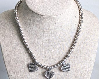 May include: A silver-toned beaded necklace featuring a series of uniform round metallic beads strung together. The necklace is accented with three dangling heart-shaped charms, each displaying a textured, vertically striped surface pattern.