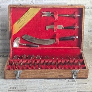 May include: A wooden box with a red velvet interior containing a set of silver cutlery. The box is labeled "Johny's Gems" and has a gold sticker with the words "Guaranteed Quality & Price" and "Factory." The cutlery includes forks, spoons, and knives.