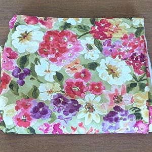 May include: A folded piece of fabric featuring a watercolor-style floral pattern. The design includes clusters of white, pink, red, and purple flowers with green leaves set against a light olive green background. The edges show a white selvage.
