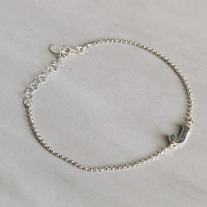 May include: A silver bracelet with a beaded chain and an adjustable clasp. The bracelet features the letter "J" at the end of the chain. The bracelet is displayed on a light gray surface.