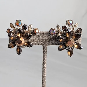May include: A pair of vintage flower-shaped earrings. The earrings feature large, dark brown faceted crystals in the center, surrounded by smaller crystals and gold-toned metal leaves. Smaller iridescent crystals add a touch of color.