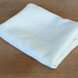 Puede incluir: Servilleta de lino plegada de color crema con un delicado diseño floral bordado. La servilleta está cuidadosamente doblada, mostrando la fina textura de la tela. El fondo es una superficie de madera cálida.