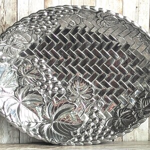 Könnte beinhalten: Ein ovales Serviertablett aus silberfarbenem Metall mit einem zentralen Korbgeflechtmuster. Der Rand ist mit geprägten Traubenclustern und großen Weinblättern verziert. Die metallische Oberfläche weist ein poliertes, reflektierendes Finish auf.