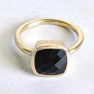 May include: A gold-toned ring featuring a square-cut, faceted black gemstone set in a smooth metal bezel. The band is thin and circular, with the hallmark 925 clearly visible on the inner surface, indicating sterling silver construction.
