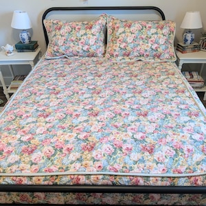 May include: A bed featuring a quilt and two matching pillow shams with a dense floral pattern in shades of pink, yellow, and blue. The bedding has a light-colored piped edge and is set on a black metal bed frame with a headboard and footboard.