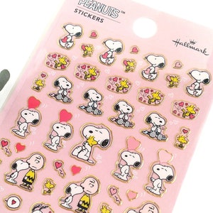 Puede incluir: Una hoja de pegatinas de Peanuts con Snoopy, Woodstock y corazones sobre un fondo rosa. Las pegatinas tienen detalles dorados y son de Hallmark. La hoja incluye el texto "PEANUTS STICKERS".