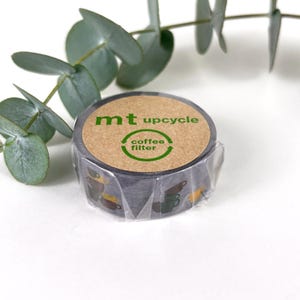 May include: A roll of washi tape featuring a dark grey background with a pattern of coffee cups in brown, green, and yellow. The label reads "mt upcycle coffee filter" in green lettering.