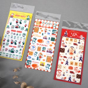 May include: Three sheets of colourful stickers featuring retro Japanese designs. The first sheet features a panda and mahjong tiles. The second sheet features a panda and the text "Mazuiya" and "Caramel". The third sheet features a postman and the text "Retro Post" and "Retro".