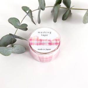 May include: A roll of masking tape with a pink and white plaid design. The tape is in a clear plastic container with the words "masking tape" and "made in Japan" printed on the label. A sprig of green leaves is in the background.