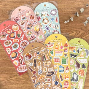 Könnte beinhalten: Eine Sammlung von Stickerbögen mit verschiedenen Themen. Die Aufkleber zeigen Essen, Blumenarrangements, Schreibwaren und Katzen. Jeder Bogen ist in einer transparenten Plastikhülle verpackt, mit einem gebogenen Oberteil und japanischem Text.