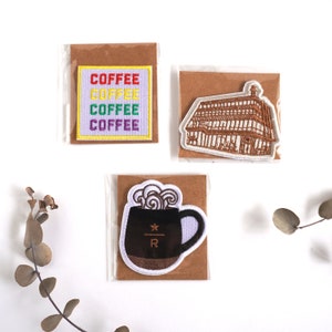 May include: Three embroidered patches on brown cards. One patch says "COFFEE" in rainbow colours. Another depicts a building with brown and white details. The third patch is a black coffee mug with steam and a star logo.