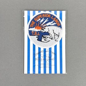 May include: A circular embroidered patch featuring a white polar bear, an orange sun, and the text THE SUPERIOR LABOR T.S.L. on a blue background. The patch is mounted on a white card with blue vertical stripes and printed text including STANDARD AND STRONG.