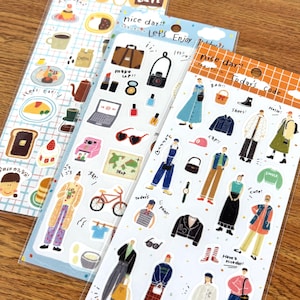 Puede incluir: Tres hojas de pegatinas con varias ilustraciones. Una hoja presenta alimentos y bebidas como café y tostadas. Otra tiene imágenes temáticas de moda y viajes, incluyendo una maleta y gafas de sol. La tercera hoja muestra ropa y accesorios.