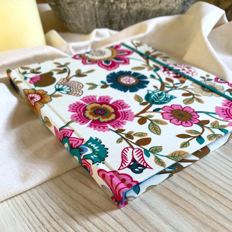 May include: A white notebook with a floral pattern in shades of pink, brown, teal, and blue. The notebook has a teal ribbon bookmark.