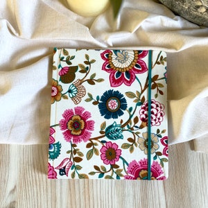 May include: A white notebook with a floral pattern in shades of pink, brown, teal, and blue. The notebook has a teal elastic closure.