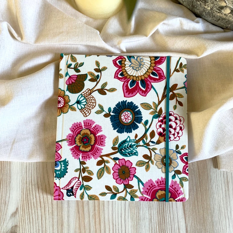 May include: A white notebook with a floral pattern in shades of pink, brown, teal, and blue. The notebook has a teal elastic closure.