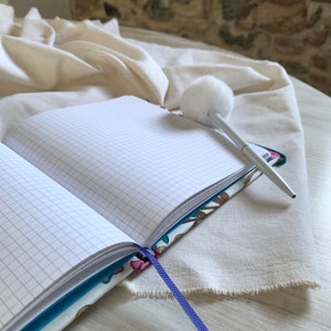 May include: A blue and white patterned notebook with a white pom-pom pen on top of it. The notebook is open and has lined pages. The notebook is on a white fabric surface.