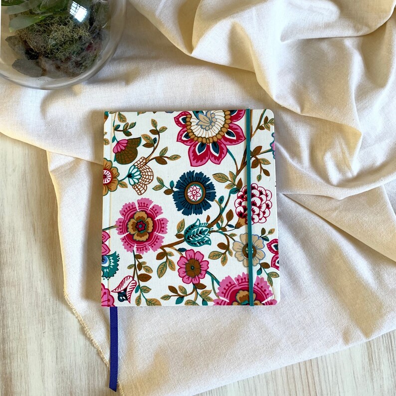 May include: A white notebook with a floral design featuring pink, teal, and brown flowers. The notebook has a purple ribbon bookmark and a teal elastic closure.