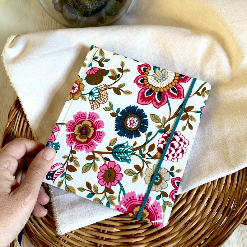 May include: A white notebook with a floral design in shades of pink, blue, green, and brown. The notebook has a teal elastic closure.